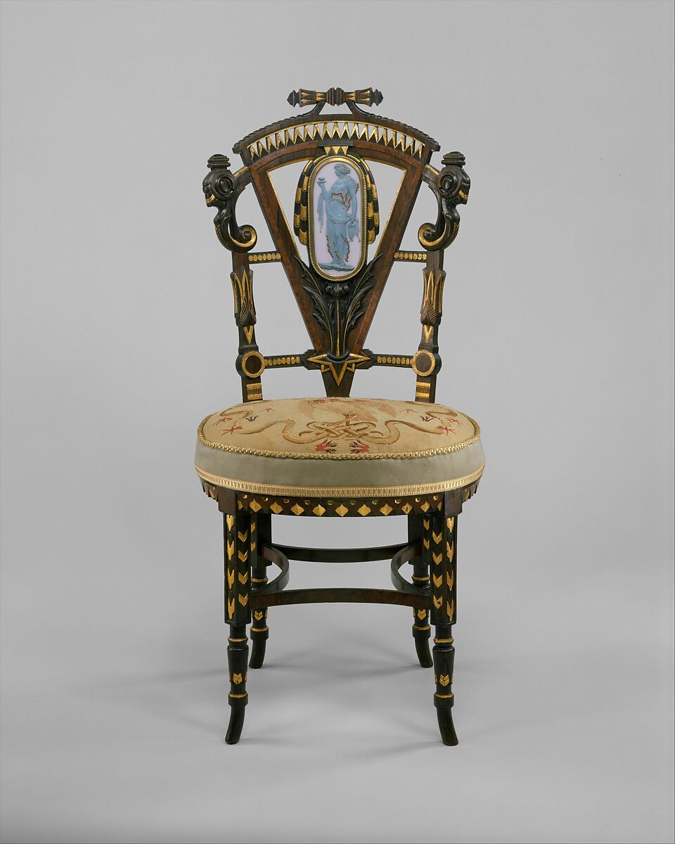 An ornate wooden side chair has gilded Egyptian motifs, a decorated back and a pale upholstered seat.