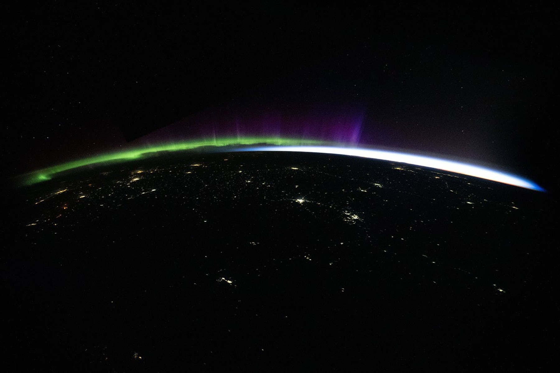 Green and purple auroral light arcs above Earth’s curved horizon, with city lights below and stars above.