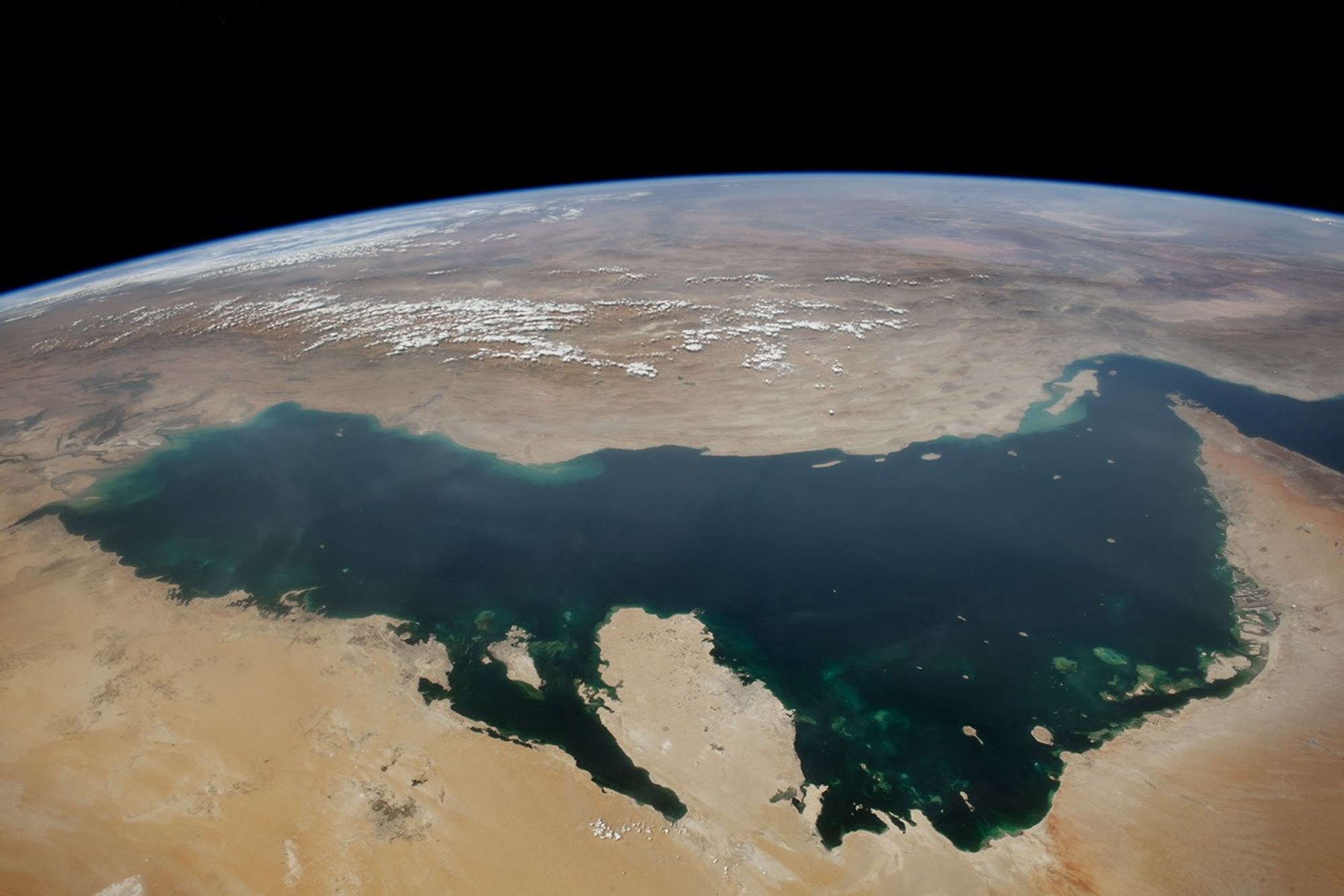 The dark blue Persian Gulf curves between desert coastlines beneath Earth’s horizon, with the Strait of Hormuz at the right.