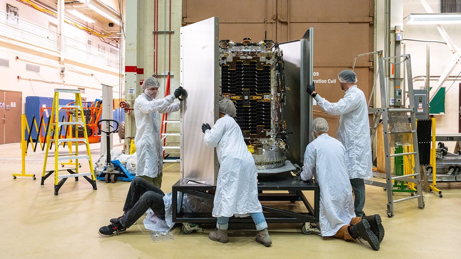 Engineers in protective clothing remove tall panels from around the upright LINK spacecraft.