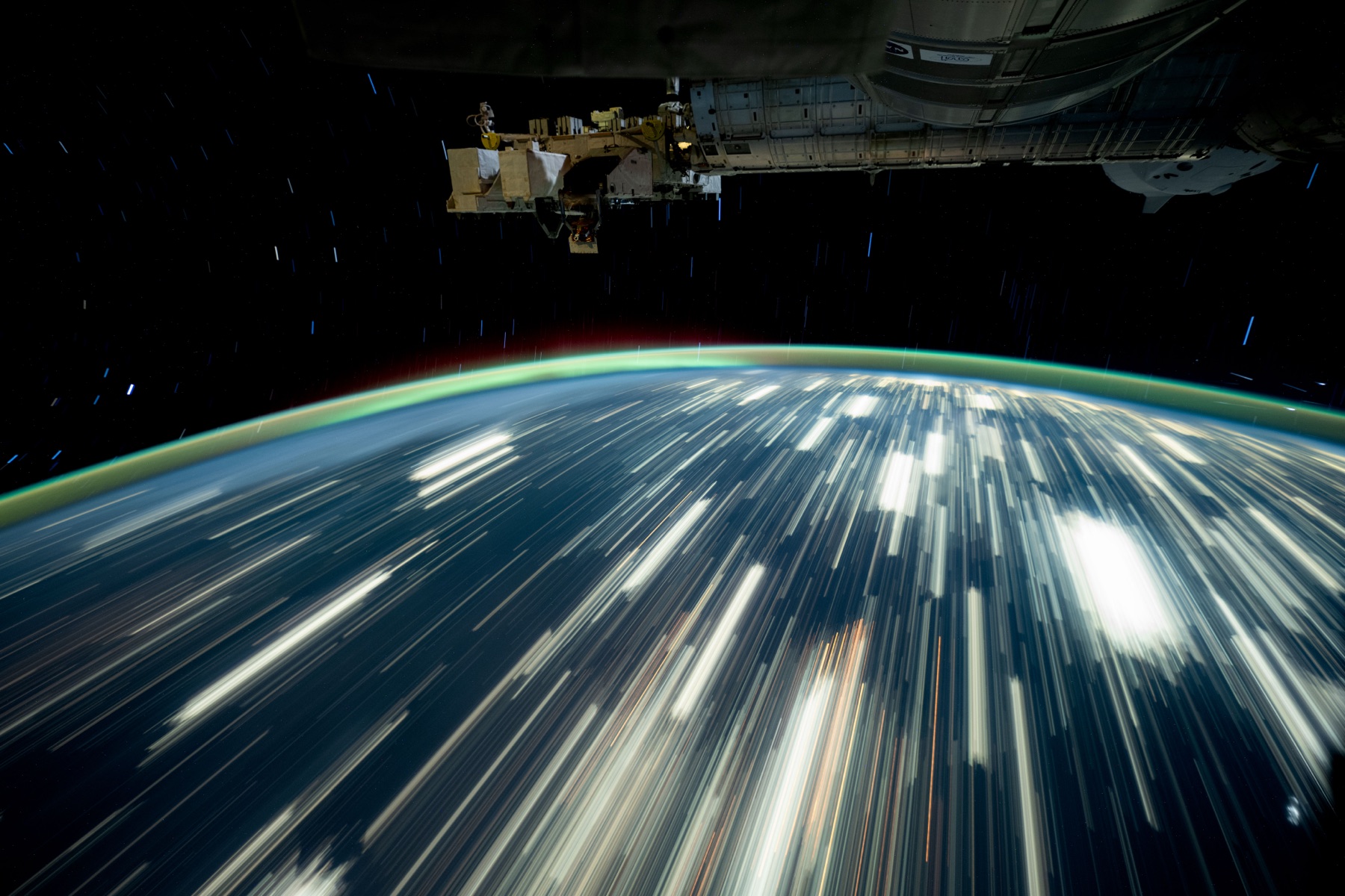 Earth’s city lights form long streaks below a green atmospheric glow, with International Space Station hardware overhead.
