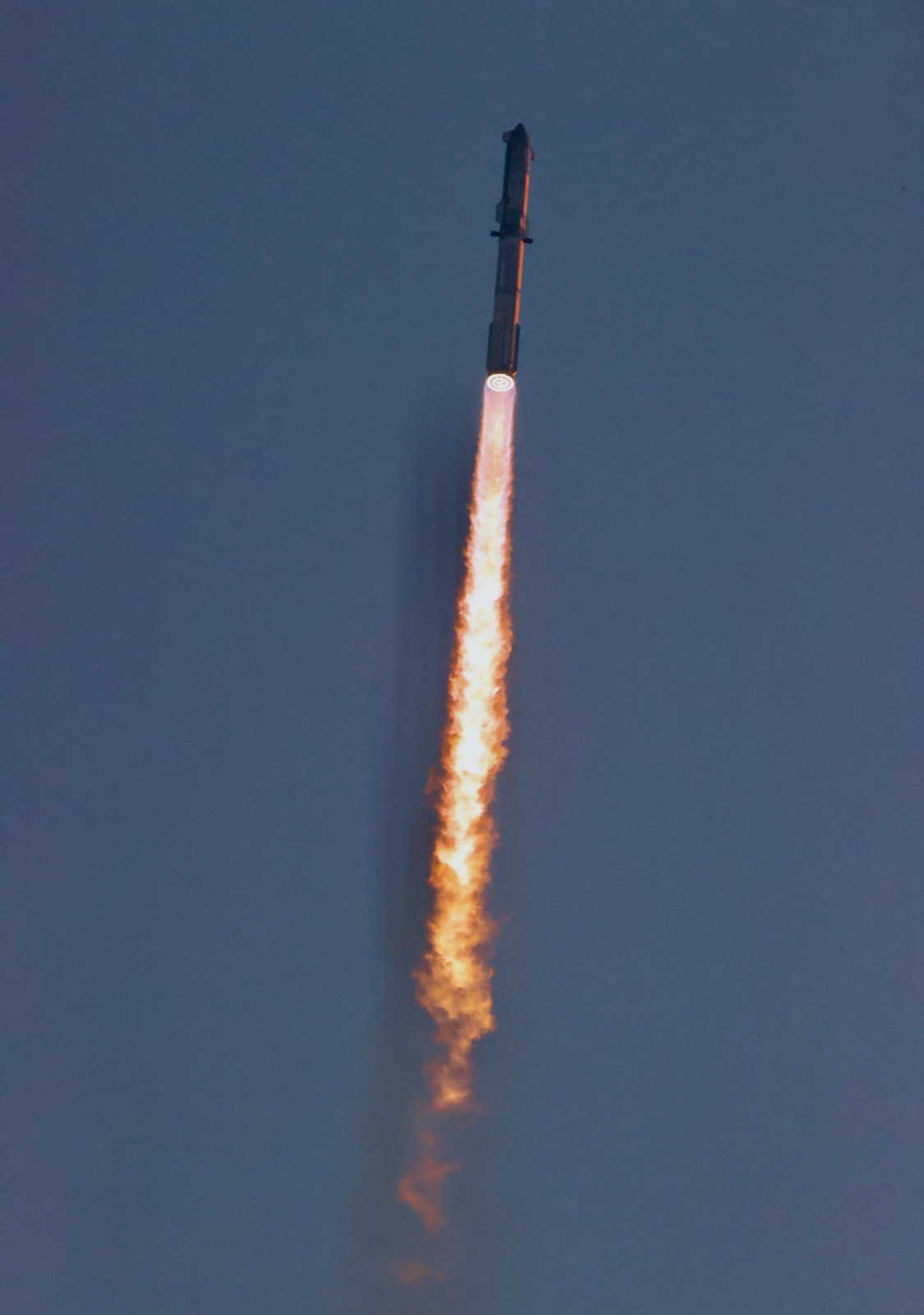 Starship climbs against a blue sky with a long orange exhaust plume beneath it.